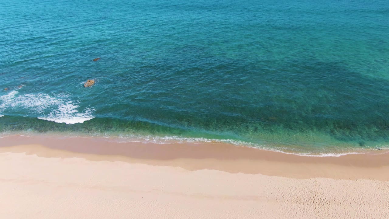 Top-down La Fortuna shoreline view with one umbrella near the lower sand edge, a horizontal line of foam, and turquoise ocean darkening farther from shore.