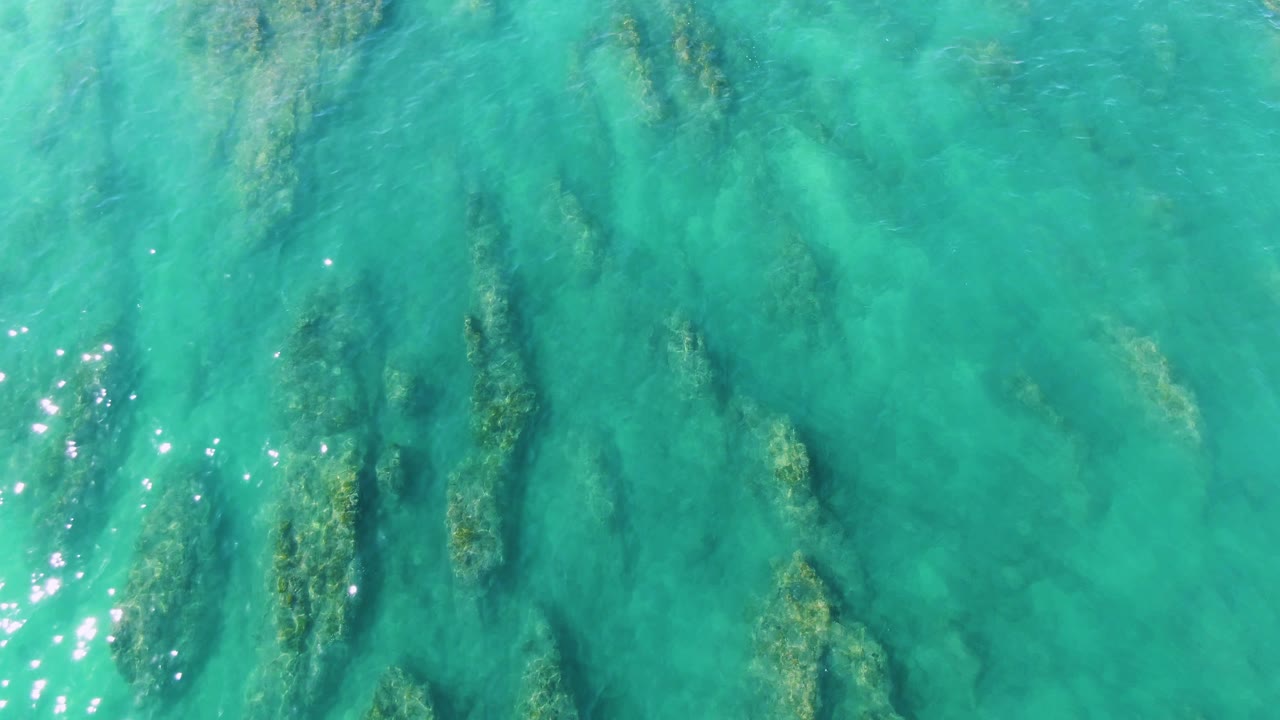 Aerial-style frame over La Fortuna turquoise water, with elongated submerged vegetation forms and scattered surface sparkles, with no visible people or structures.