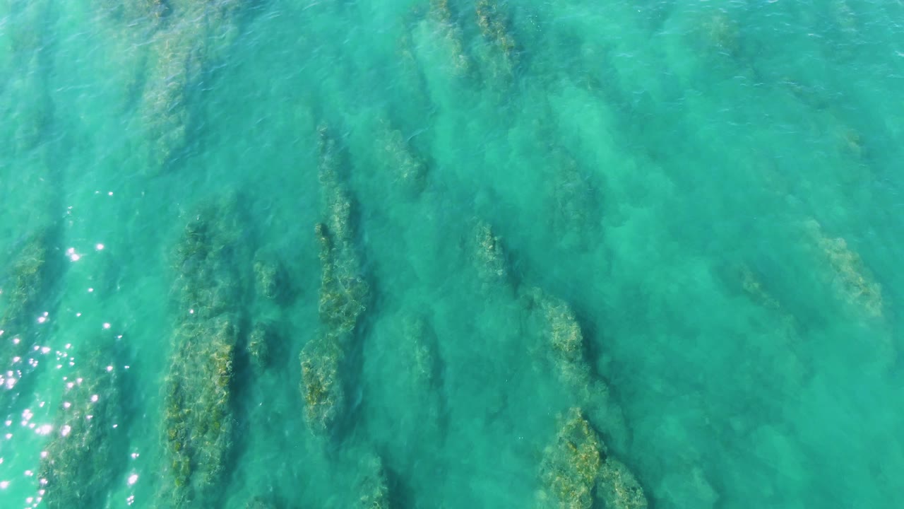 Blue-green water at La Fortuna shows submerged plant forms below a gently rippled surface, with a cluster of bright reflections along the lower left and no visible people.