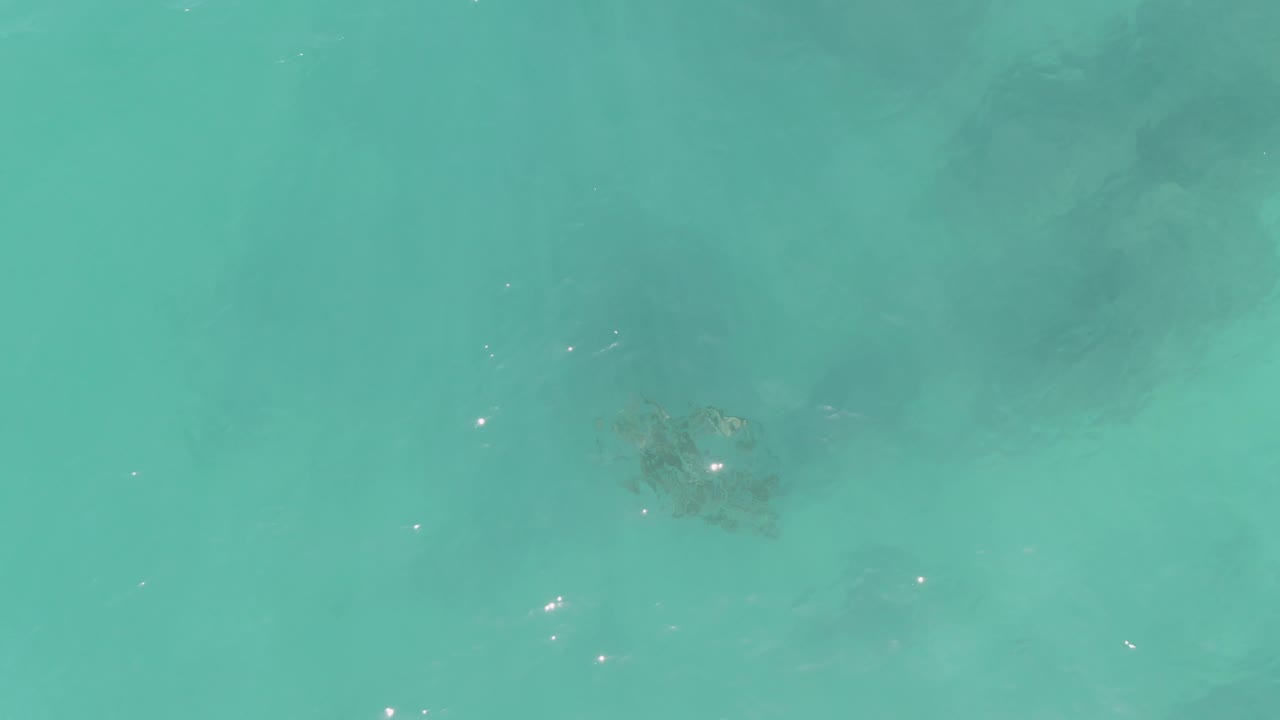 Clear turquoise water at La Fortuna, Baja California Sur, with a small dark underwater formation slightly left of center and faint sun glints across the surface, with no visible people.