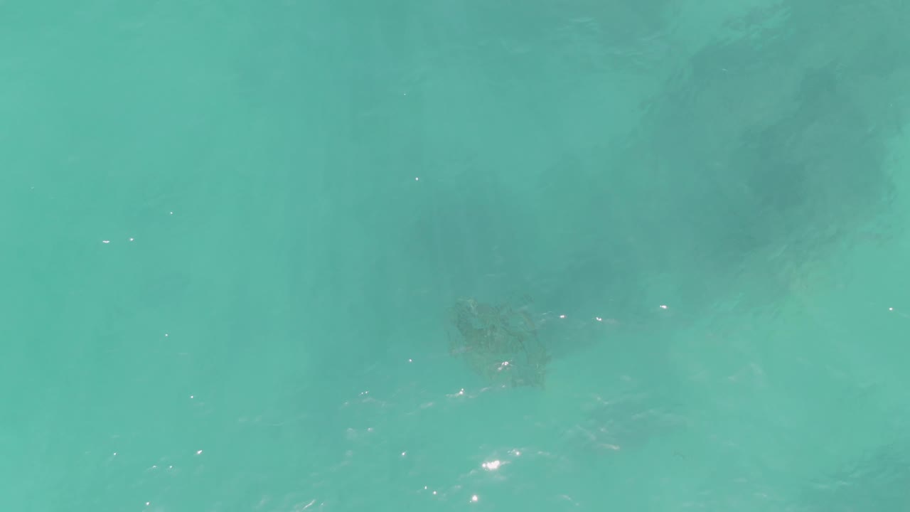 Aerial view over turquoise ocean at La Fortuna, Baja California Sur, with darker submerged rocks near the right side and soft ripples on the water, with no visible people.