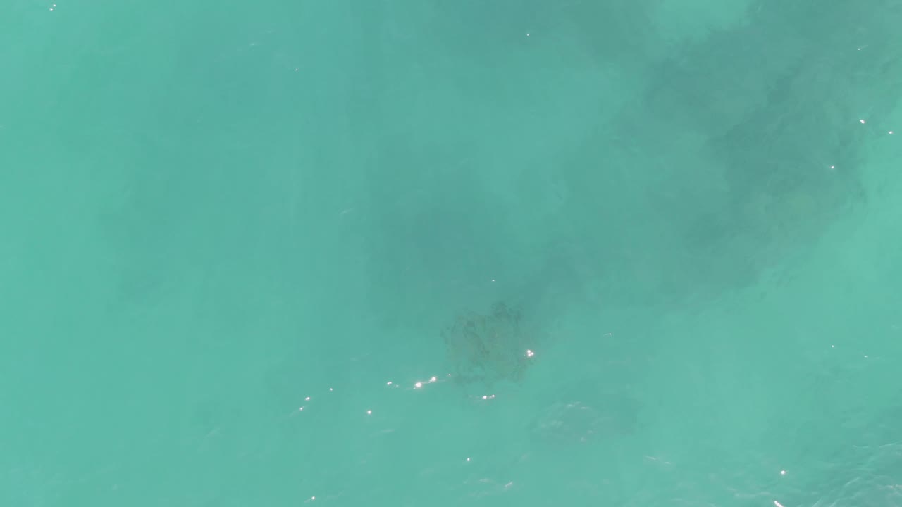 Top-down turquoise water at La Fortuna, Baja California Sur, with shadowy underwater patches and a few bright surface reflections near the lower left, with no visible people.