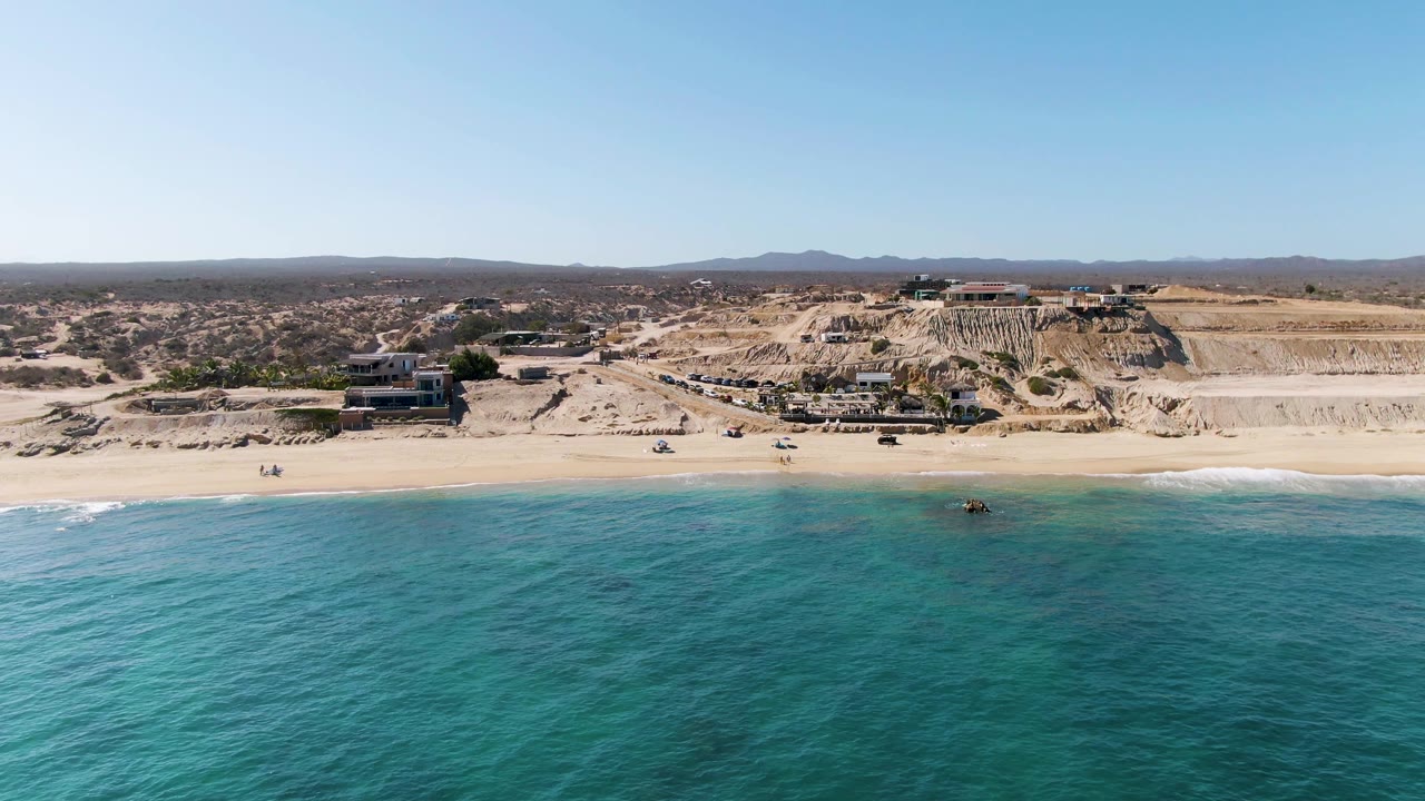 Wide aerial frame of La Fortuna, Baja California Sur, with turquoise water in front, a sandy beach, cliffside buildings, dirt roads, vehicles, and tiny beachgoers visible.