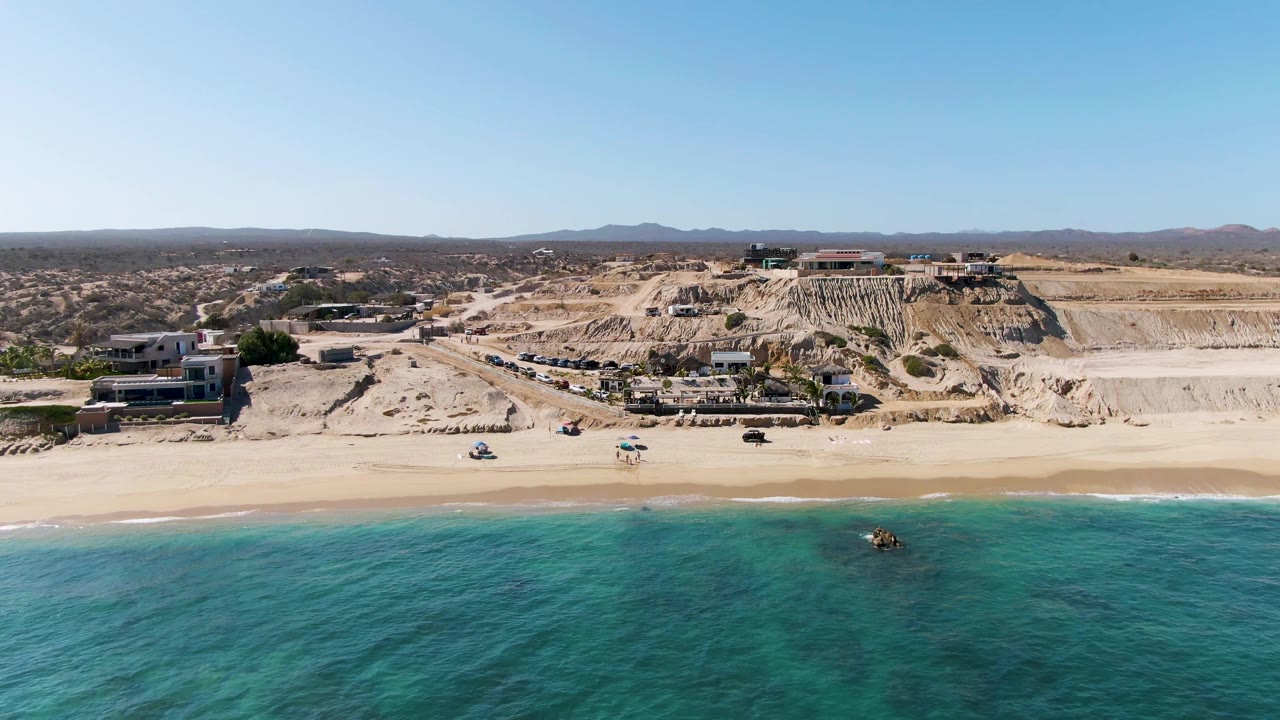 Aerial frame over La Fortuna, Baja California Sur with turquoise water in the foreground, pale sand, dry cliffside buildings, and no visible people at the start of the clip.