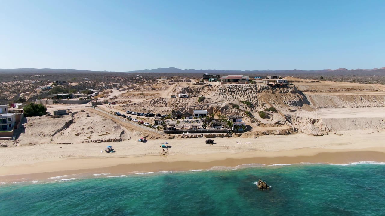 Aerial frame in La Fortuna, Baja California Sur with the golf cart farther along the shoreline, a few beach umbrellas, clear water, dry cut cliffs, and no visible people.