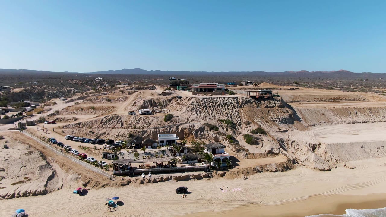 Opening aerial frame over a La Fortuna desert quarry with terraced sand cuts, a parking row, low buildings, distant hills, clear blue sky, and no visible people.
