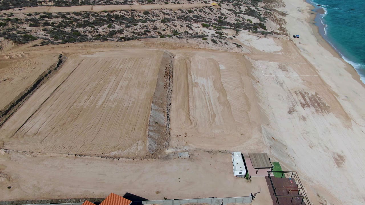 Drone view over a graded sandy beach in La Fortuna, Baja California Sur, with turquoise ocean at right, scrubby dunes inland, a small white vehicle near the lower edge, and no visible people.