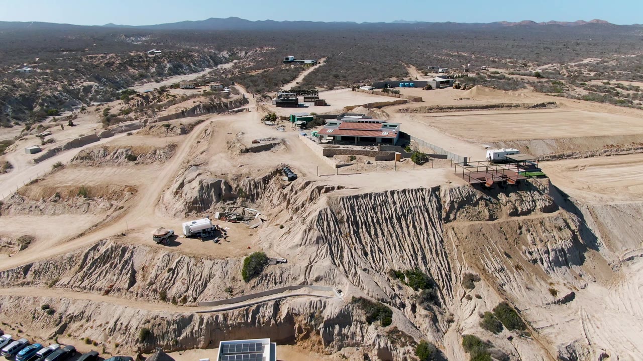 Aerial frame over a sandy excavation site in La Fortuna, Baja California Sur, with red-roofed buildings, graded roads, cut earth banks, distant hills, and no visible people.