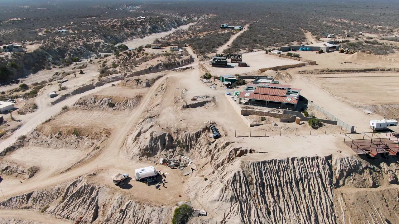 Aerial perspective over the La Fortuna excavation site, showing ridged exposed earth in the foreground, red-roofed buildings to the right, desert tracks, and no visible people.
