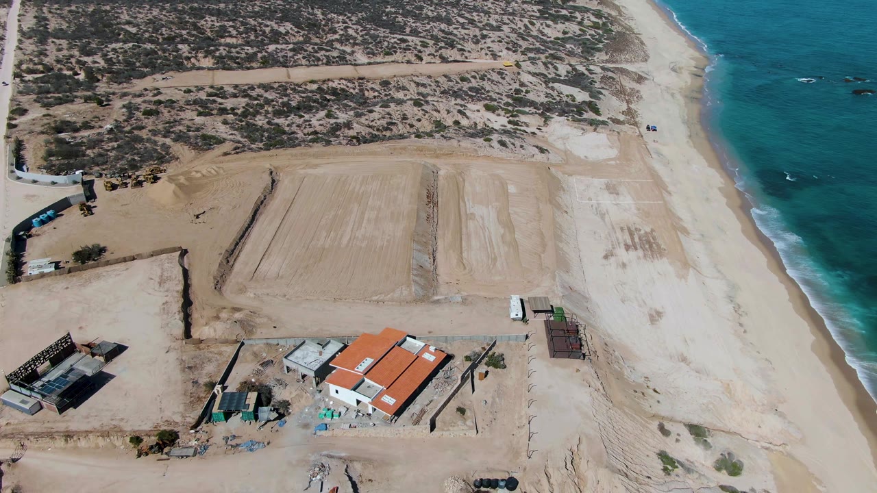 Aerial frame over La Fortuna, Baja California Sur showing a sandy beach, turquoise ocean at right, dry scrubland inland, a coastal road, red-roofed construction buildings, and no visible people.