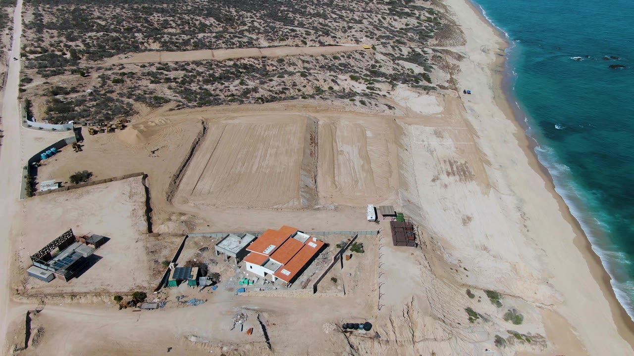 Aerial frame from La Fortuna, Baja California Sur with the white vehicle slightly farther along the coastal road, red-roofed buildings below center, pale sand, blue water, and no visible people.