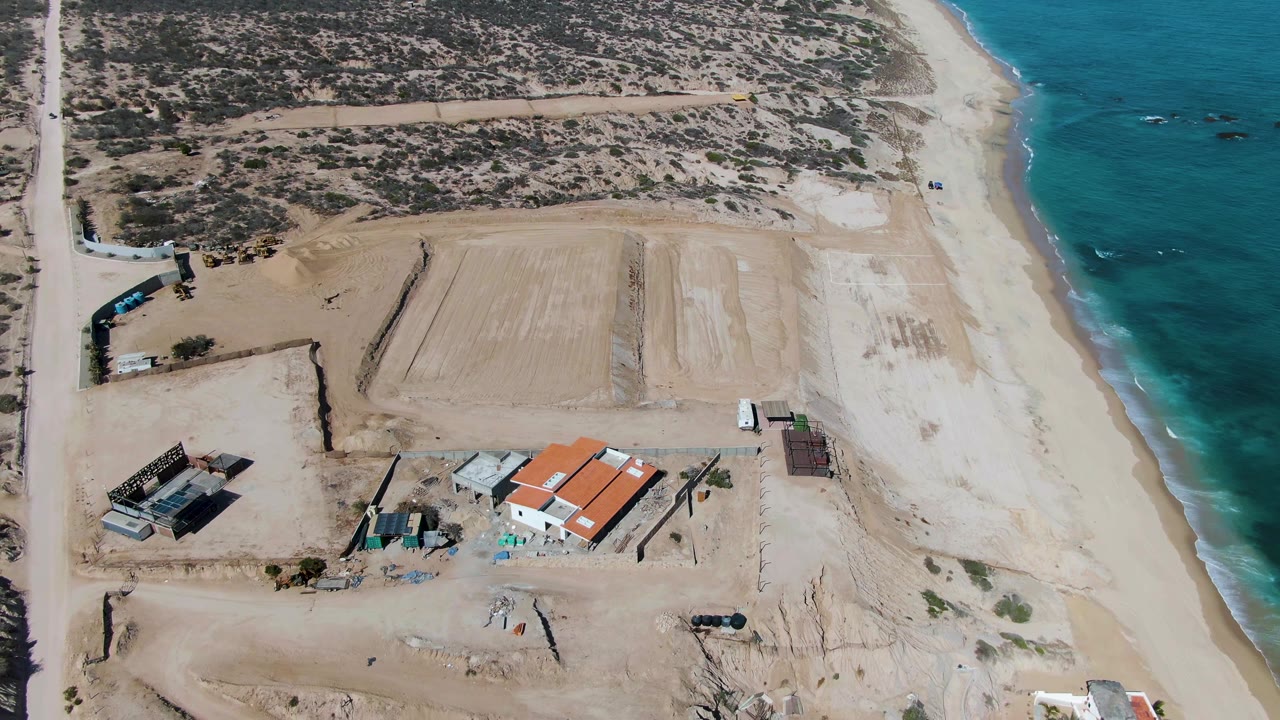 Aerial frame over La Fortuna, Baja California Sur with scrubland at top, a rectangular graded sandy area near the beach, red-roofed construction buildings near the lower center, ocean surf at right, and no visible people.