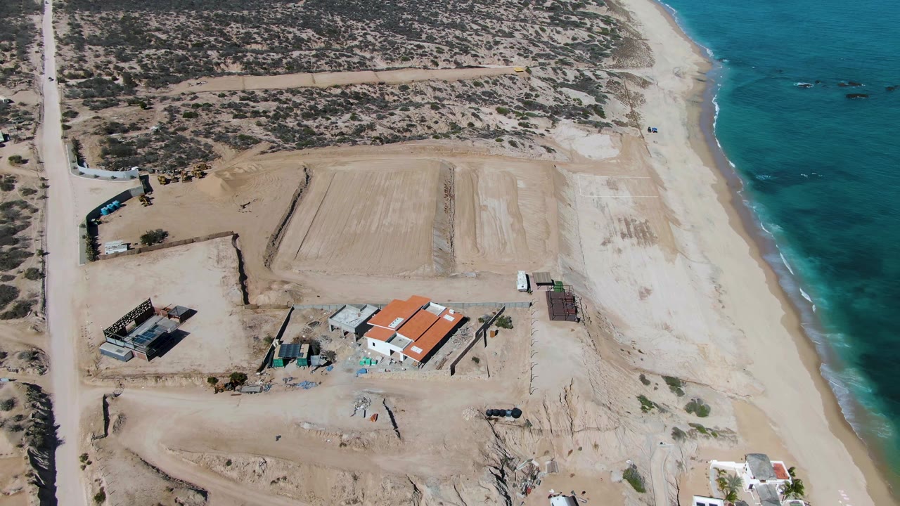 High aerial view of a beach road, dry scrub, construction buildings, and turquoise sea.