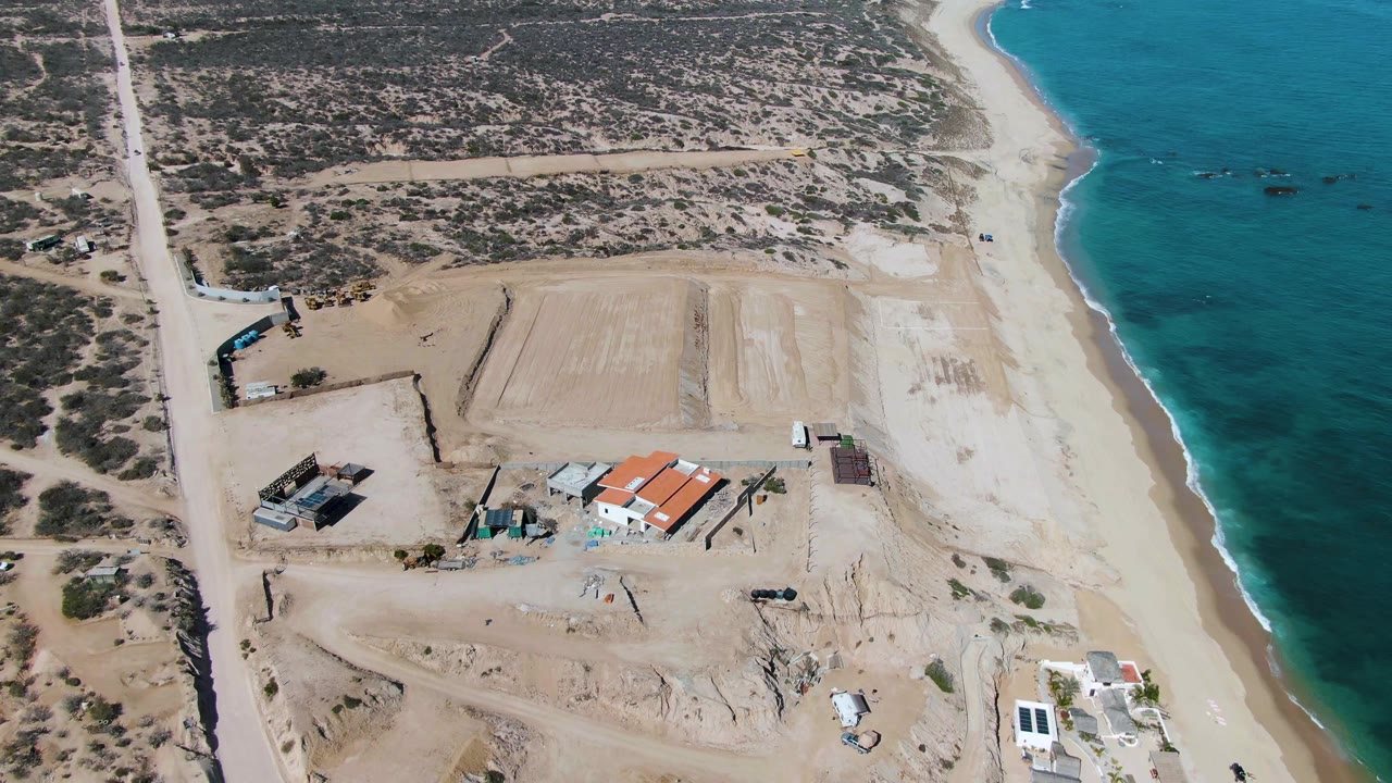 Aerial frame over La Fortuna, Baja California Sur showing a sandy construction site, a straight dirt road, low buildings, turquoise shoreline, a small vehicle near the lower left, and no visible people.