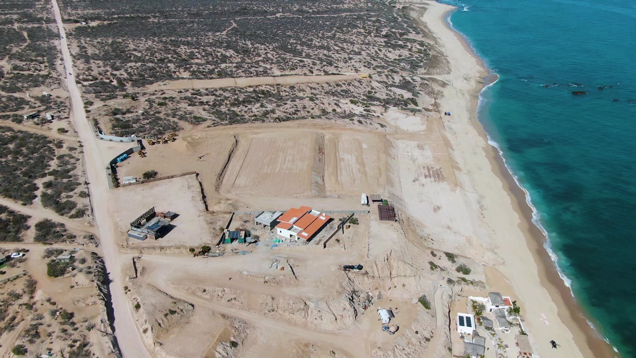 Aerial frame over La Fortuna, Baja California Sur with the road along the left edge, graded sand and buildings in the center, turquoise water to the right, and no visible people.
