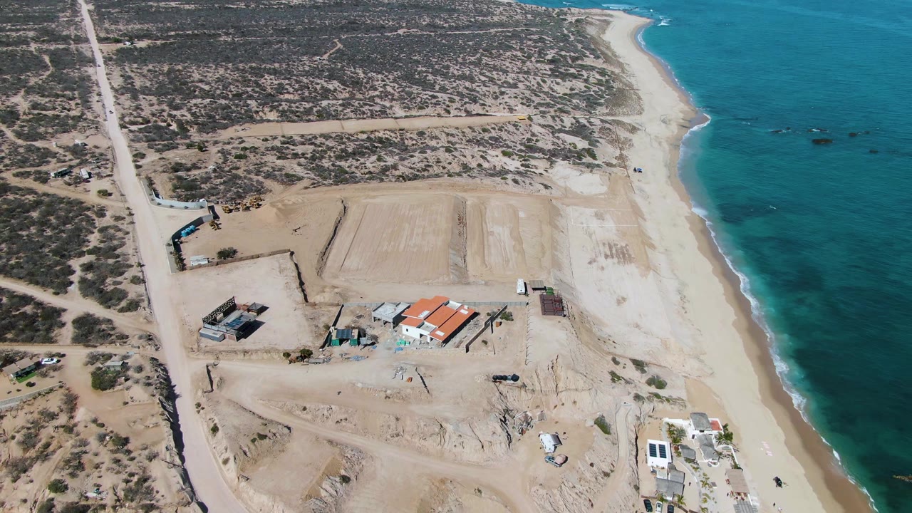 Aerial frame over La Fortuna, Baja California Sur showing dry scrubland, a pale sandy work area, scattered structures, a narrow beach, turquoise ocean, and no visible people.