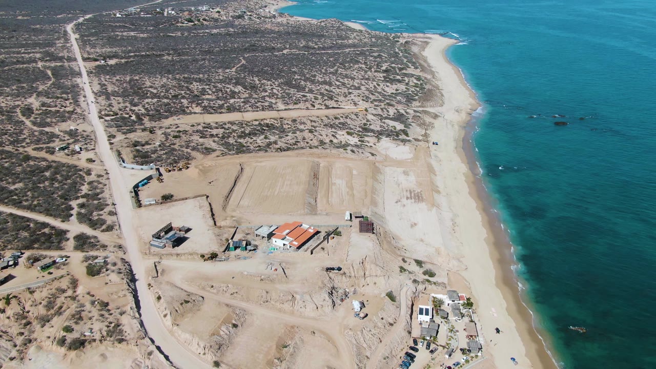 Opening aerial frame over La Fortuna, Baja California Sur, showing pale beach, turquoise ocean, desert scrub, roads, sand piles, small structures, and tiny people near the shoreline.
