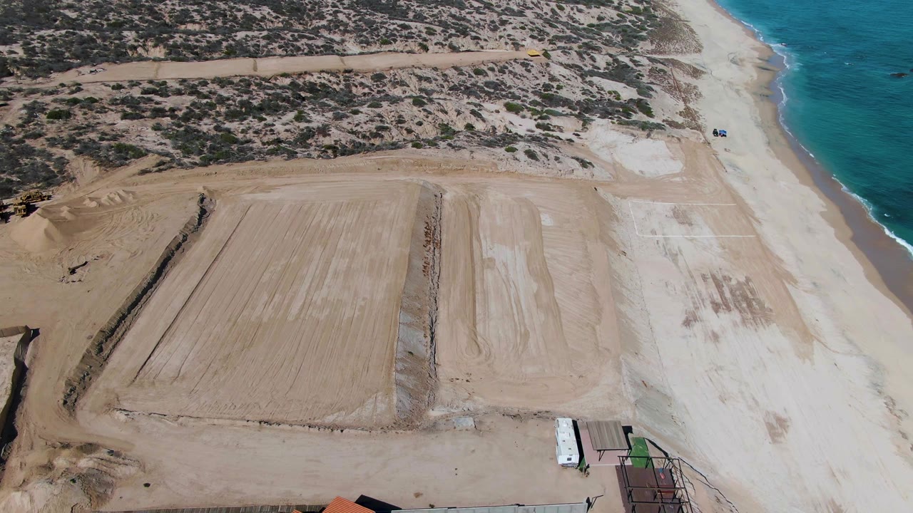 Aerial frame over La Fortuna, Baja California Sur, showing a graded sandy construction site beside the beach and blue ocean, with no visible people.