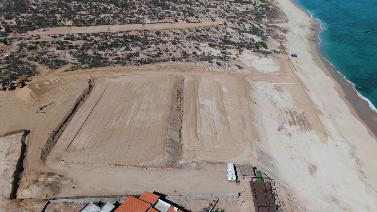 Aerial view in La Fortuna, Baja California Sur, with rectangular sandy pads centered, the ocean on the right, and small roofed structures beginning to enter the lower edge, with no visible people.