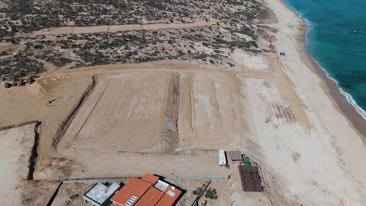 Aerial frame from La Fortuna, Baja California Sur, showing sandy development ground, a pale beach, blue water, and red-roofed buildings more clearly along the lower edge, with no visible people.