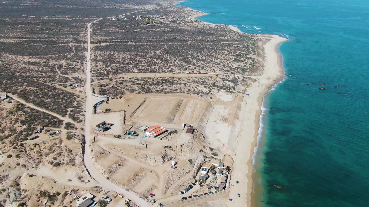 Aerial frame in La Fortuna showing beachside earthworks, dirt road access, scrubland inland, and blue ocean along the right side, no visible people.