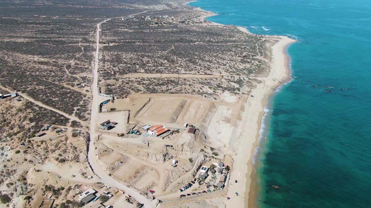 Drone-style aerial view over La Fortuna with sandy graded lots, scattered vehicles, construction machinery, coastal scrub, and turquoise water, no visible people.