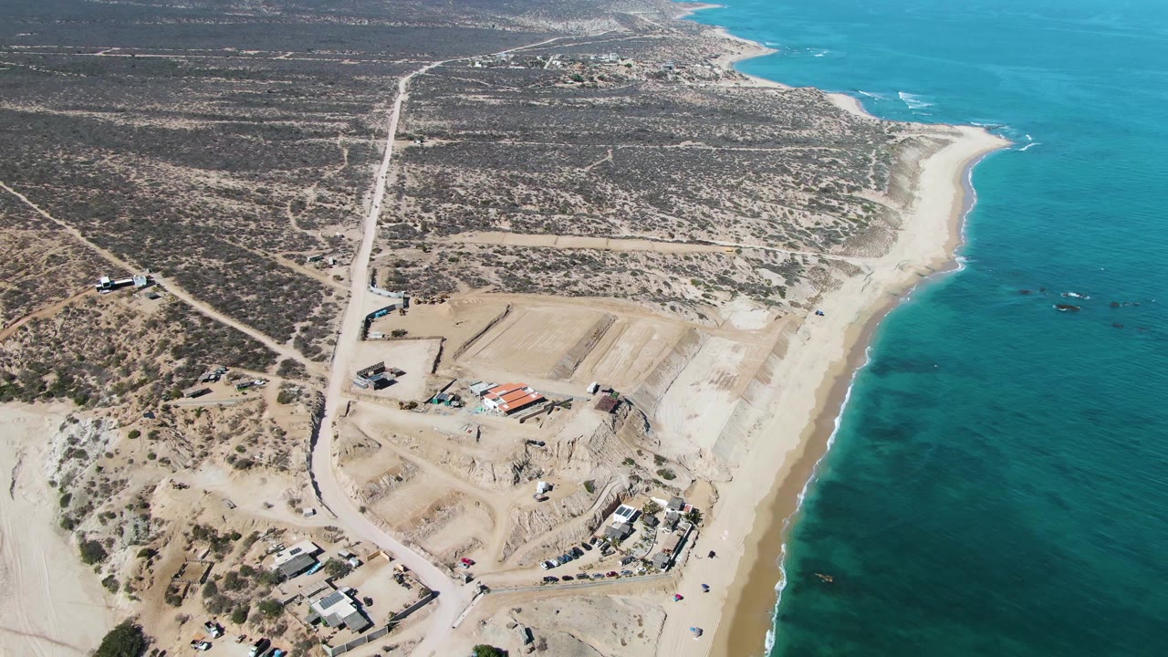 At 0:00, a high aerial view over La Fortuna shows turquoise ocean on the right, a pale sandy beach, desert scrubland, dirt roads, and a construction site with no visible people.
