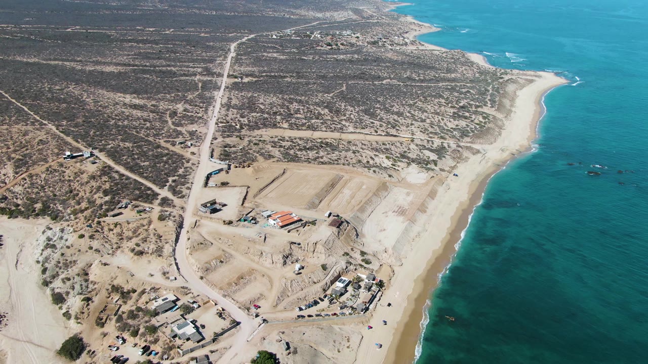 At 0:01, the aerial frame holds the La Fortuna beach and blue-green water, with the graded construction area and straight dirt road more centered inland, and no visible people.