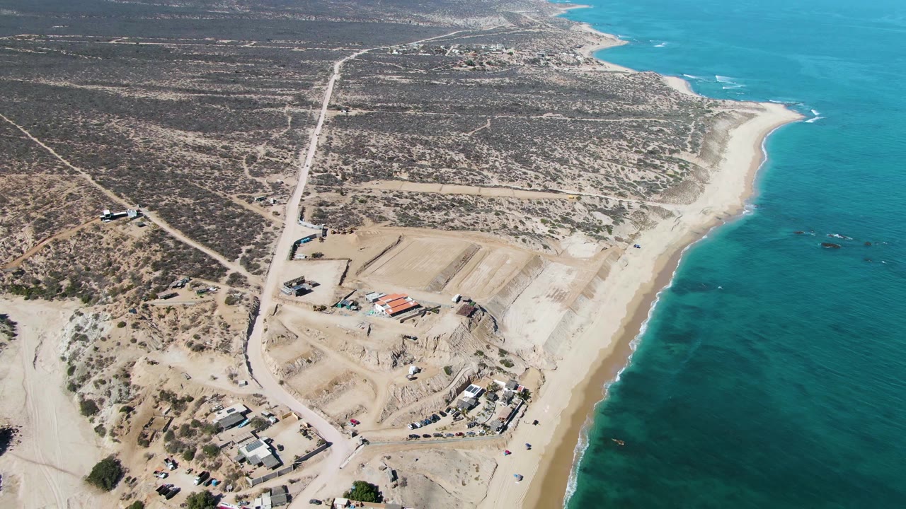 At 0:02, the La Fortuna coastline remains in a wide drone view, with the ocean along the right edge, exposed sand at the waterline, a construction zone below center, and no visible people.