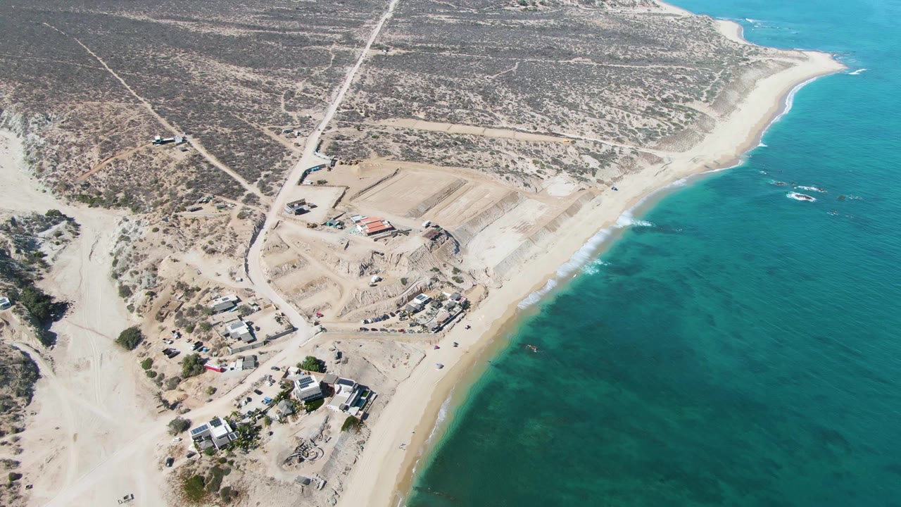 Opening aerial frame over La Fortuna, Baja California Sur showing dirt roads, graded construction areas, scattered buildings, a curved sandy beach, turquoise ocean, and no visible people.
