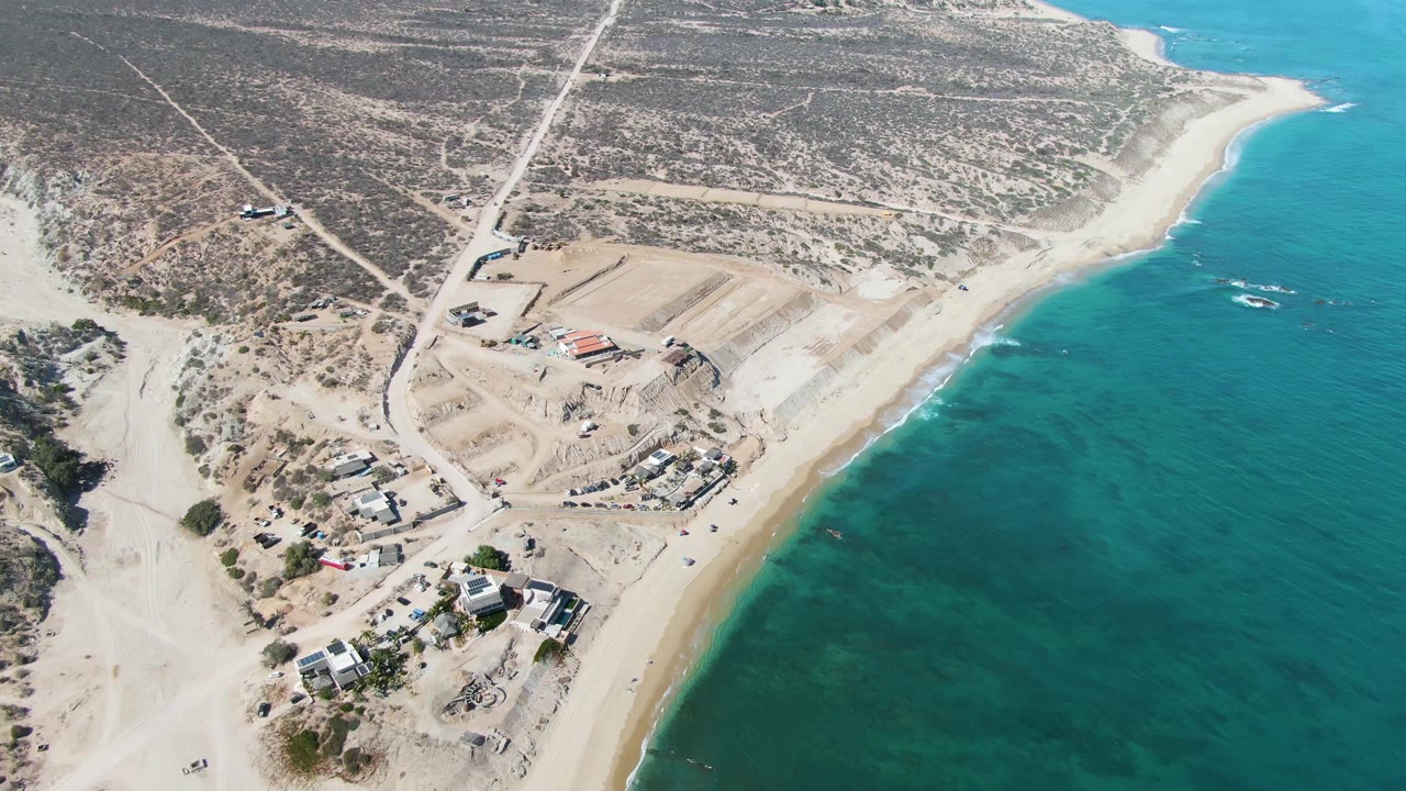 Mid-clip aerial view of La Fortuna showing a red vehicle near the road network, graded earth around unfinished structures, open desert scrub inland, bright turquoise water, and no visible people.