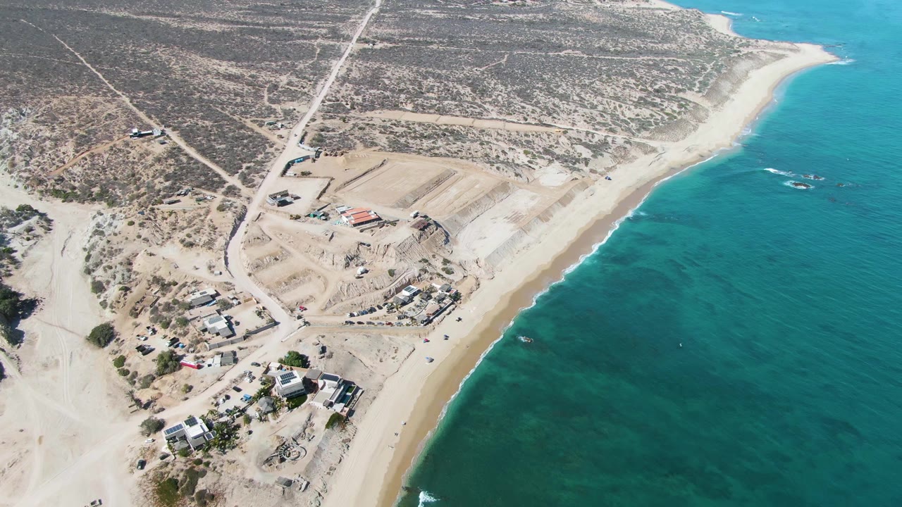 Aerial frame over La Fortuna, Baja California Sur showing a sandy shoreline, turquoise ocean, dirt roads, graded construction pads, scattered buildings, and no visible people.