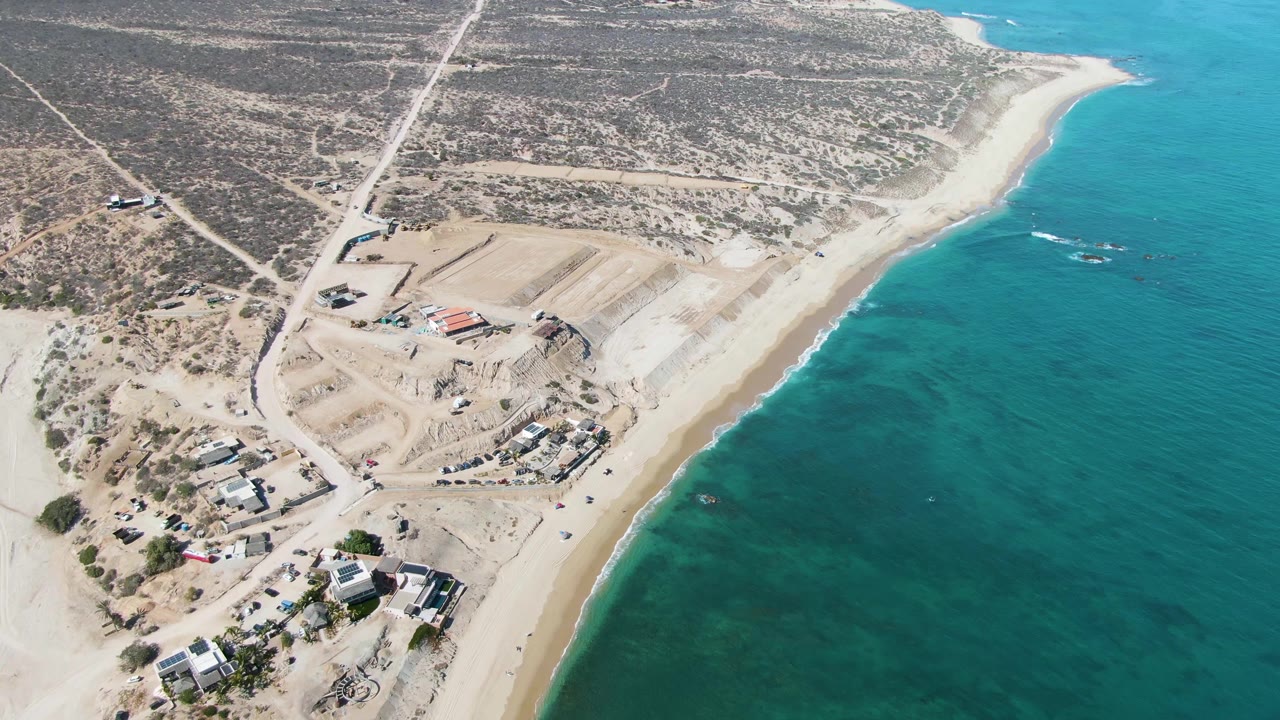 Drone perspective over La Fortuna coastline showing graded sand lots, a cluster of buildings near the lower left, a narrow beach, blue-green ocean, and no visible people.