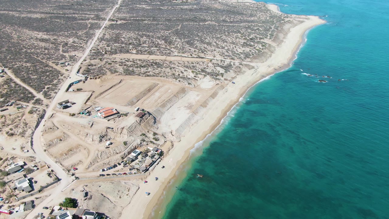 Drone view over La Fortuna with the graded construction area centered lower in frame, dirt roads cutting through dry land, clear blue-green ocean on the right, and no visible people.