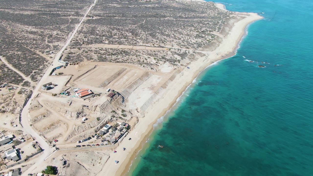 Oblique aerial over La Fortuna showing a coastal road, pale sand construction pads, small structures, a beach strip, turquoise water, and no visible people.