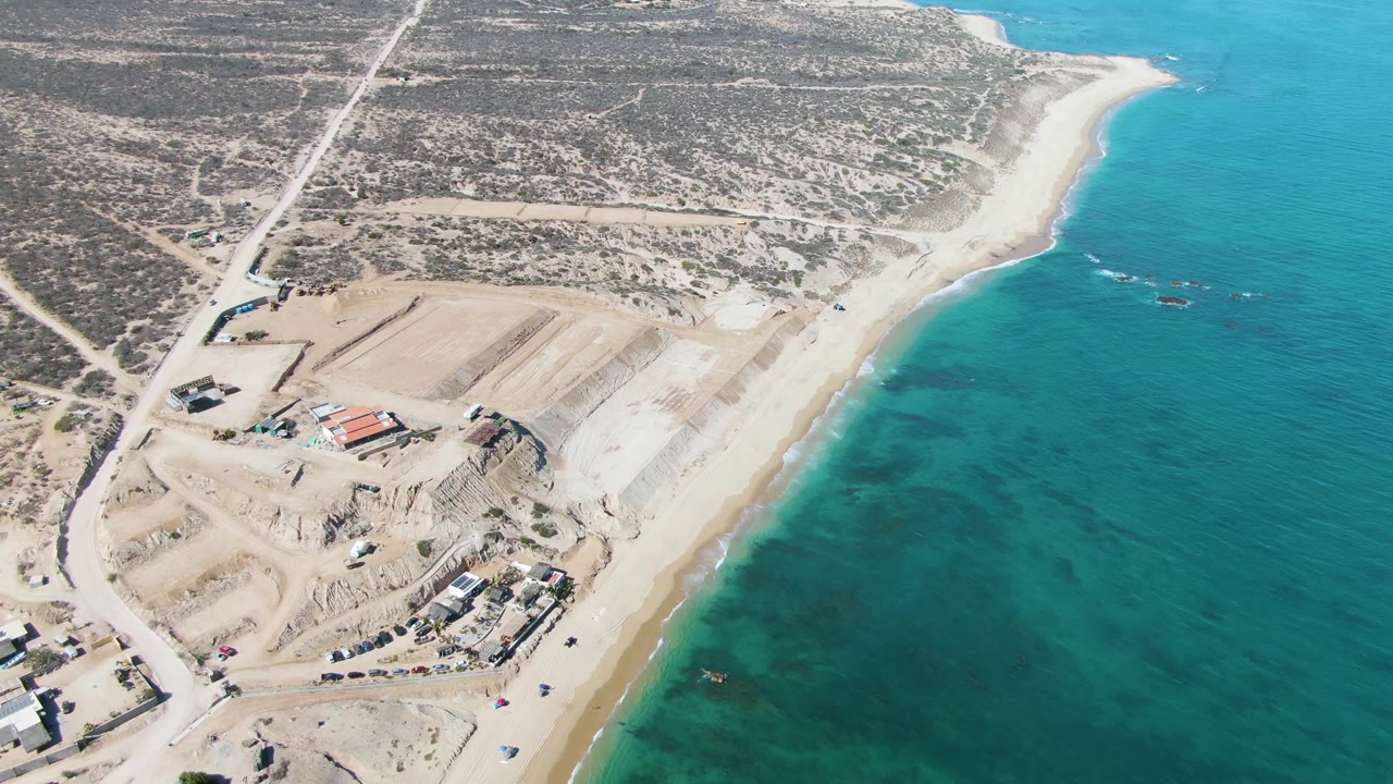Aerial frame over La Fortuna with earthworks and access roads on dry sand, a narrow beach below them, shallow turquoise sea to the right, and no visible people.