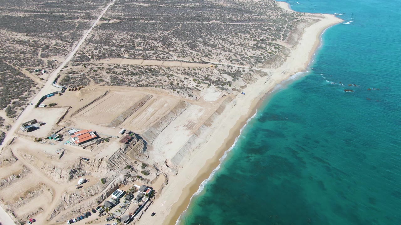 Aerial view in La Fortuna, Baja California Sur with the shoreline running diagonally, construction pads and material piles left of the beach, and turquoise ocean to the right with no visible people.