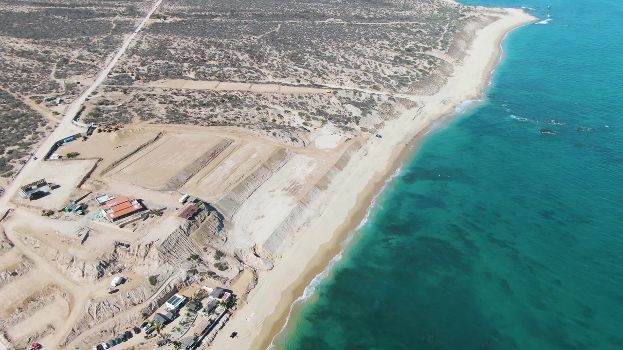 La Fortuna aerial frame showing a coastal road and dry scrubland above a sandy beach, with graded construction areas and turquoise water filling the right side, no visible people.