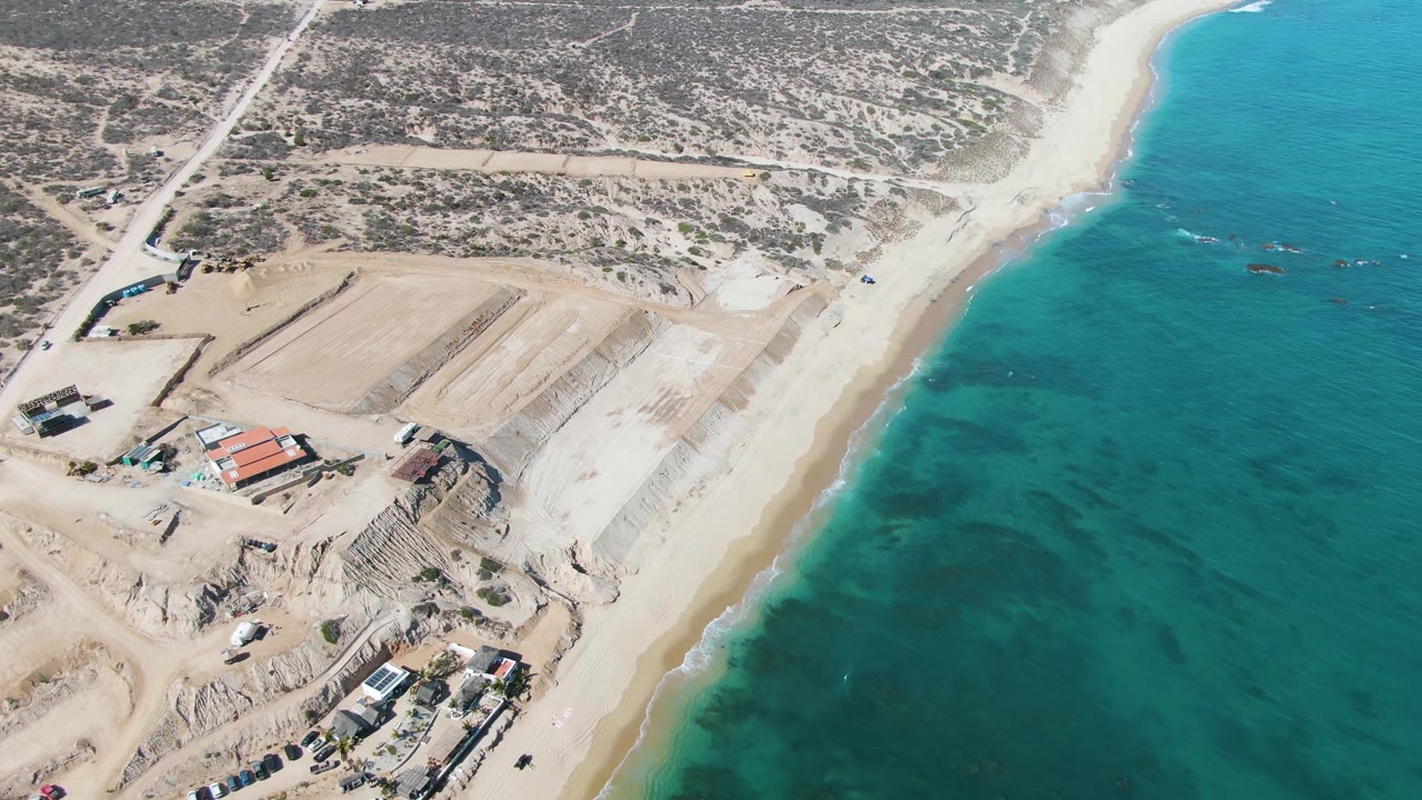 Oblique drone frame over La Fortuna with beach and light surf between blue-green water and a construction area with earth cuts, vehicles, and low buildings, no visible people.