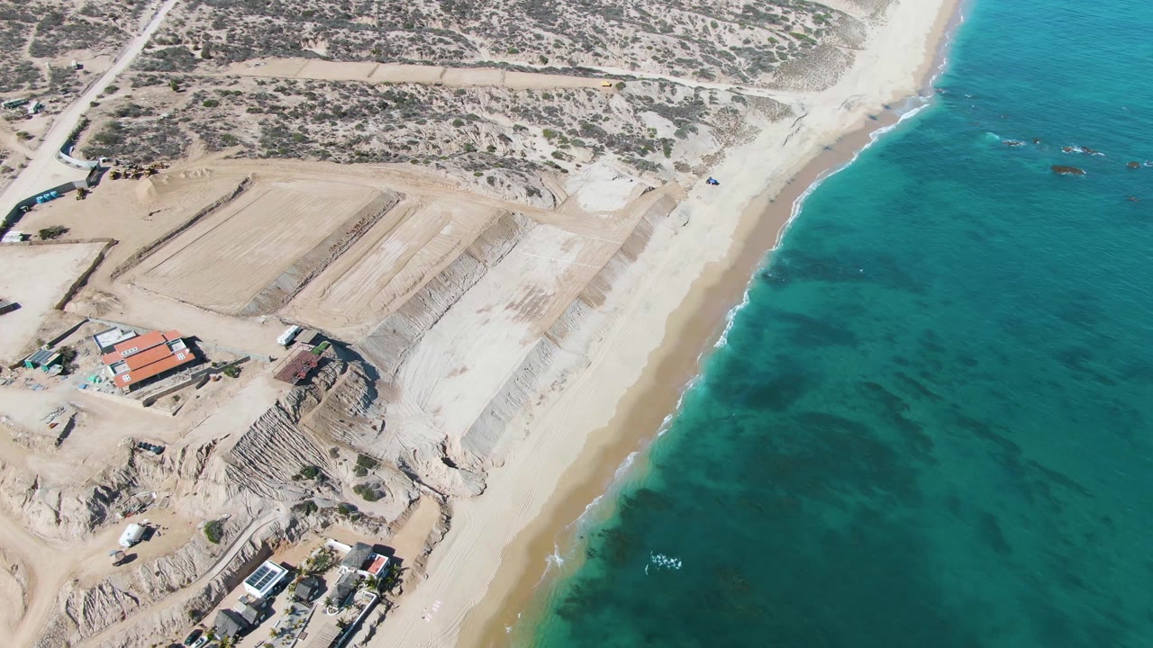 Aerial frame over La Fortuna, Baja California Sur, showing graded coastal land, small buildings, pale sand, turquoise water, and no visible people at the start of the clip.