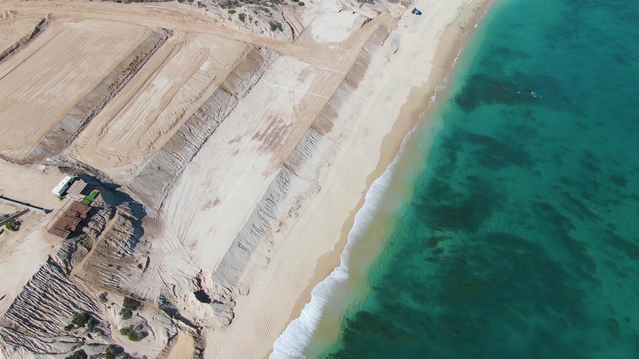 At two seconds, the overhead view shows the diagonal surf line, shallow blue-green water with darker patches, pale sand work inland, and no visible people.