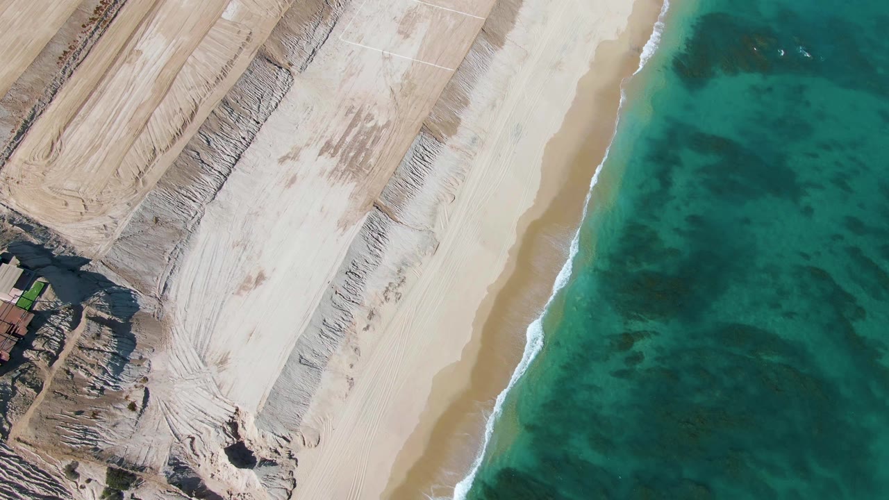 Near-vertical aerial view of La Fortuna beach with scraped pale sand, coastal cuts, a small green vehicle and structures at left, turquoise ocean at right, and no visible people.