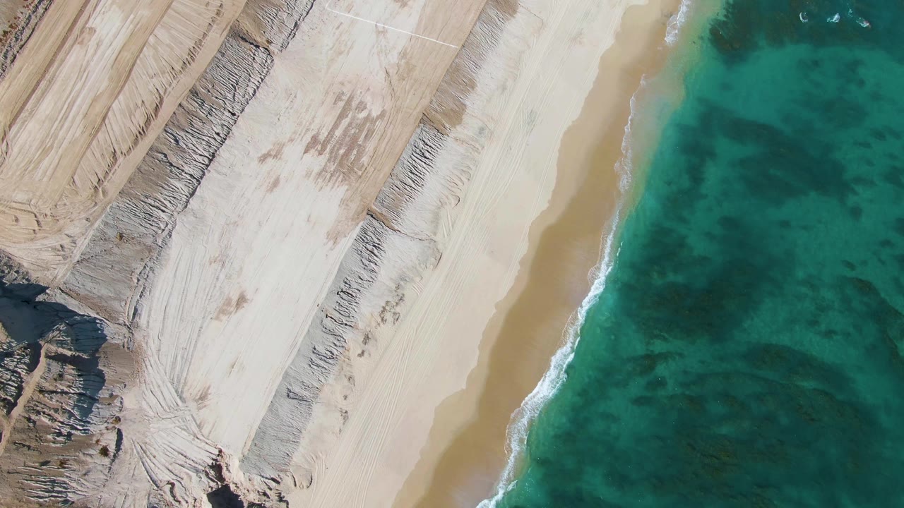 Top-down La Fortuna beach view with broad pale sand, darker scraped construction marks, layered coastal cuts on the left, turquoise ocean on the right, and no visible people.