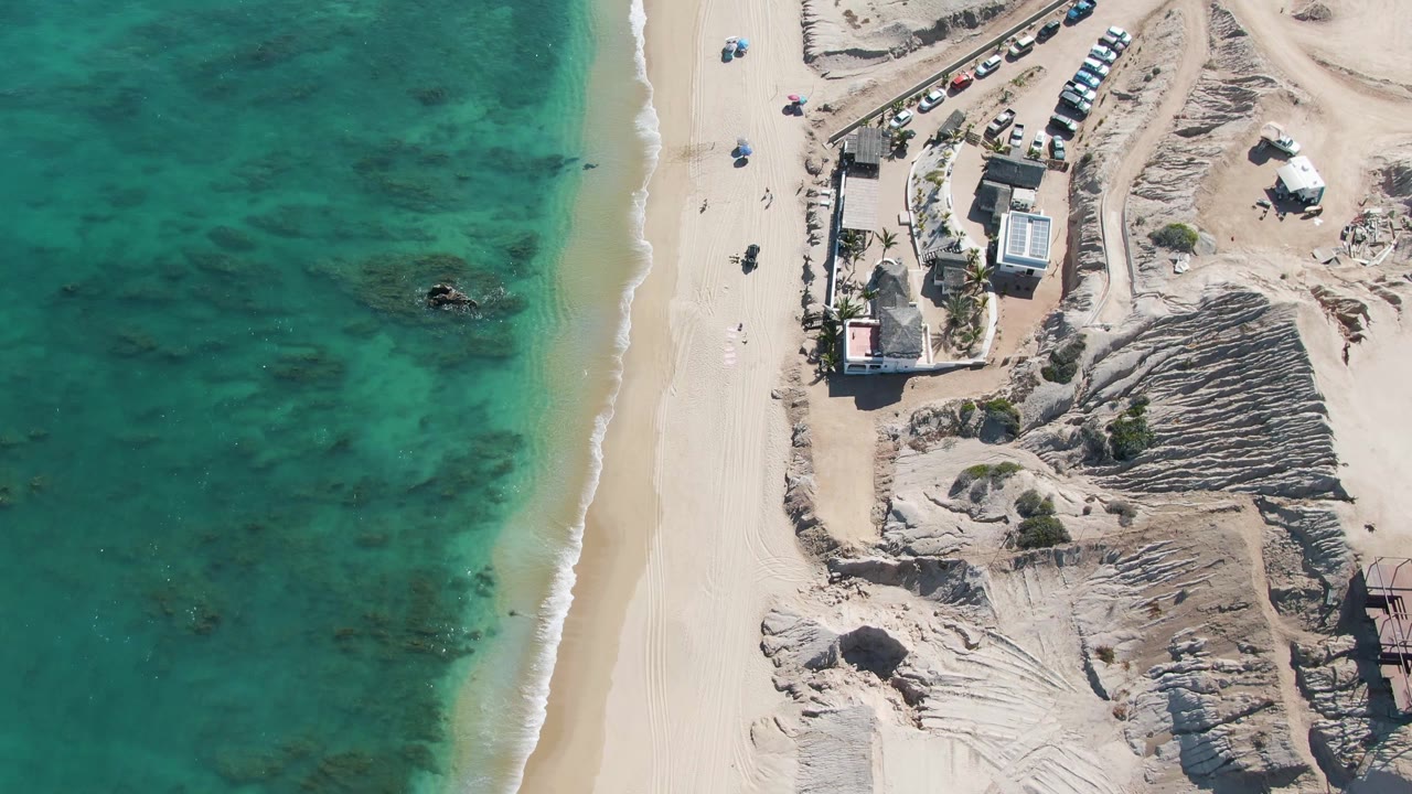 Overhead frame from La Fortuna, Baja California Sur showing turquoise water beside a pale sand beach, low buildings, parked cars, sandy tracks, and no visible people.
