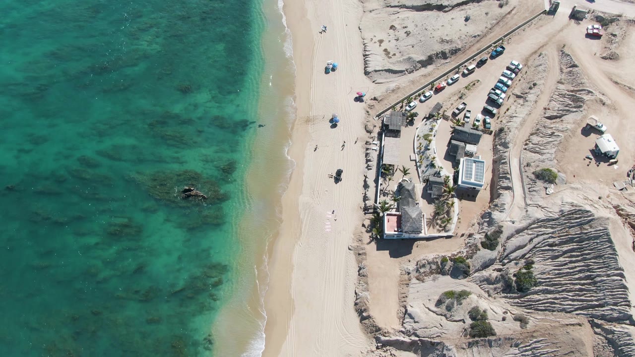 Drone-height frame over La Fortuna showing shallow turquoise water on the left, a bright sand beach in the center, and low coastal buildings with parked cars on the right, with no visible people.