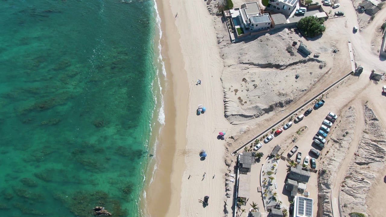 Overhead drone frame of La Fortuna, Baja California Sur with turquoise water, a narrow surf line, pale sand, beach umbrellas, parked cars, and low oceanfront buildings.