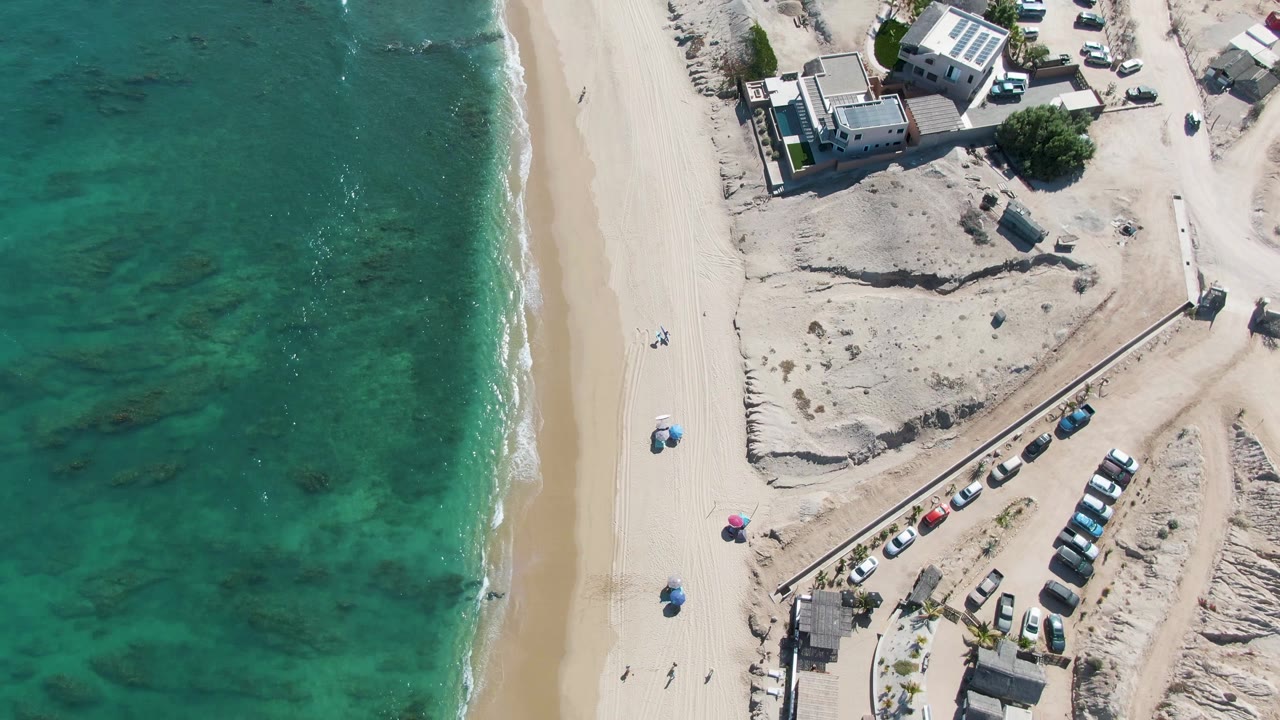 Aerial beach view with clear water, sand, umbrellas, cars, and buildings.