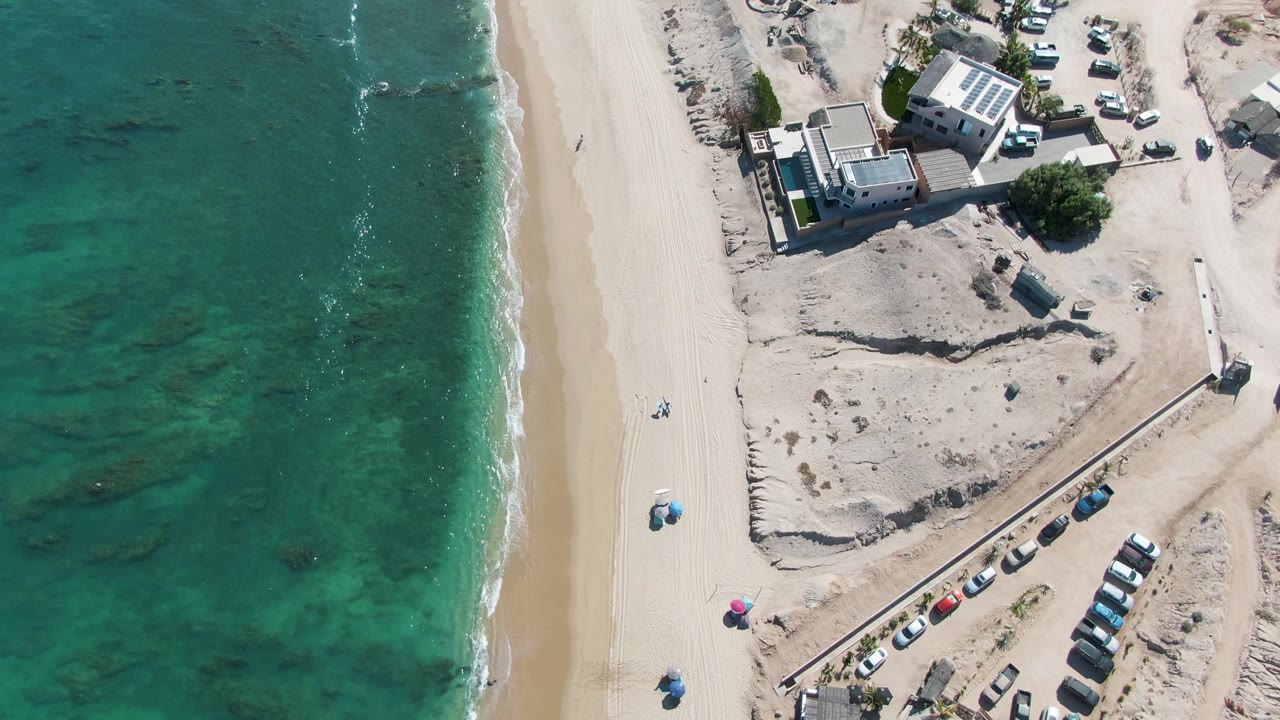 Top-down frame from La Fortuna showing turquoise ocean on the left, a clean strip of sand, small figures near umbrellas, and a cluster of buildings and parked cars beyond the beach.