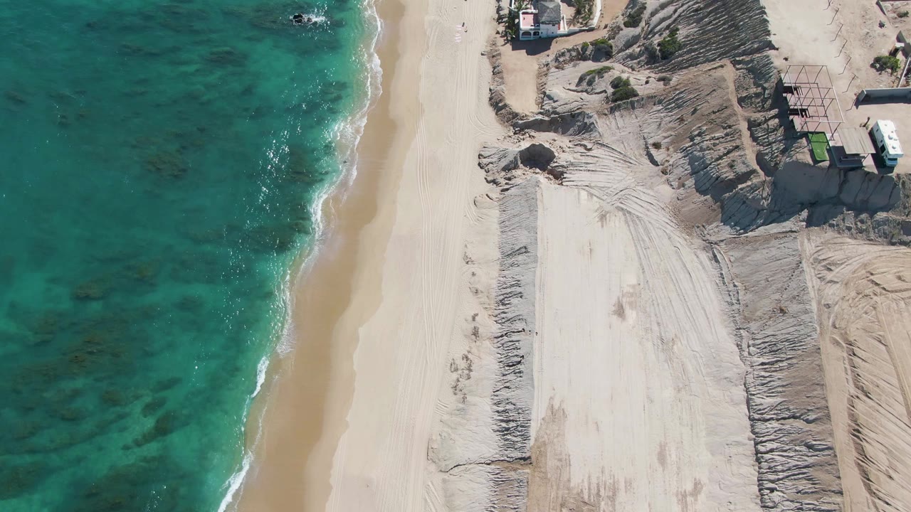 Overhead drone frame in La Fortuna, Baja California Sur, showing turquoise water at left, a pale beach path, dune cuts and small beachfront structures at right, with no visible people.