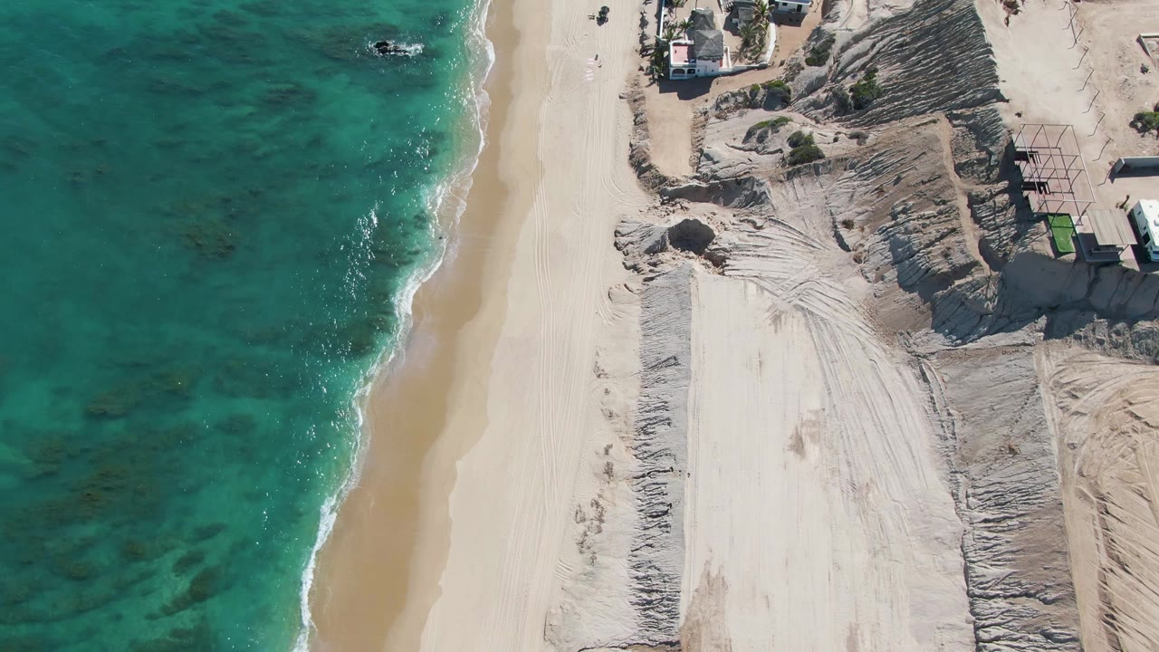 A high aerial view in La Fortuna, Baja California Sur, frames the surf line, wide sand path, dune textures, small structures, and tiny vehicles on the shore road, with no visible people.